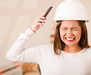 Young woman wearing construction helmet facing camera, simulating smashing mobile phone to her own head in frustration