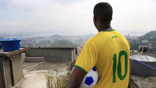 A kid in a soccer jersey stands with a ball under his arm while looking out at the city of Rio de Janeiro