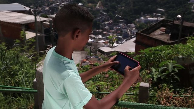 A boy stands outside and plays with his tablet while looking out over the city
