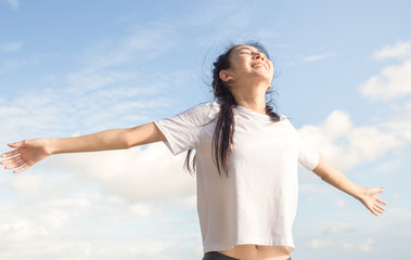 Fototapeta na wymiar Happy relaxed asian woman breathing deep fresh air and raising arms on the beach with blue sky background