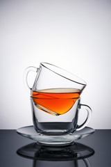 Two transparent glass cups . In the upper Cup of tea. The Cup and saucer. The Studio photos. The rear light. The reflection on the table. On 
the black table. White background.
