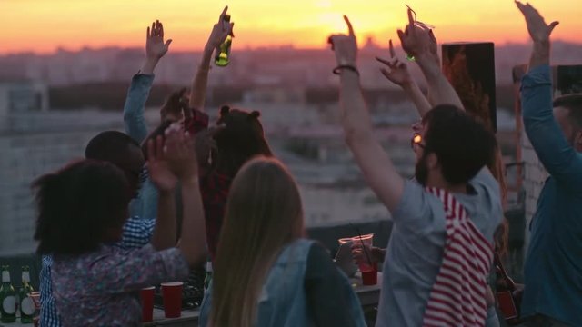 Group of multi-ethnic young people dancing and raising their arms up in air to the music played by dj at sunset urban party on rooftop