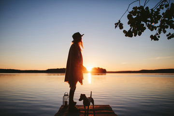 girl in a poncho and a hat with lamp standing back to the view of the sunset on the lake