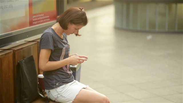 young woman sitting in waiting on the bench in the railway station, holding smartphone, looking at screen, using cell phone app, typing or reading text message
