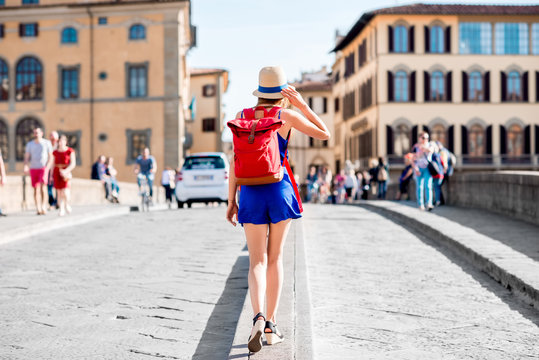 Beautiful female traveler in blue dress, hat and backpack walking on crowded Holy Trinity bridge in Florence city. Back view