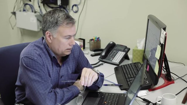 A mature stern looking network administrator or technician in office reviews data on two computer screens.