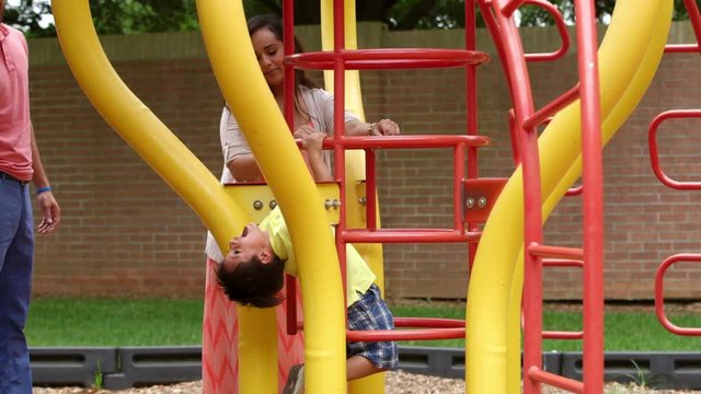 A young boy hangs upside down on a playground monkey bars while mom looks on