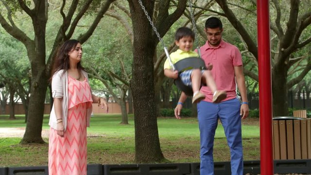 A mom and dad spend some quality time with their little son swinging him on a playground swing