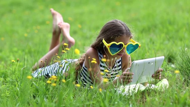 Beautiful girl in big sunglasses with tablet computer lies on the grass. Girl playing the game on his tablet computer. Little girl in glasses lies on the green grass and uses tablet pc