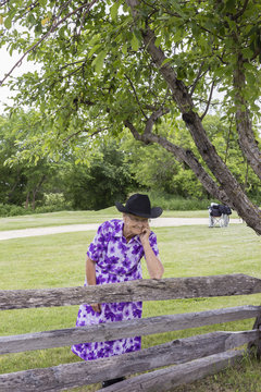 vertical image of an elderly lady acting comical wearing a cowboy hat and leaning against the fence beside a big green tree in summer.