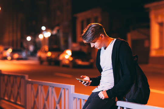 Young Man Reading Smartphone Outside. Attractive Guy With Bag Sitting On Street Fence With Phone. Technology Addiction Concept