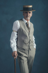 Retro 1920s dandy in suit standing with cane in front of gray wa