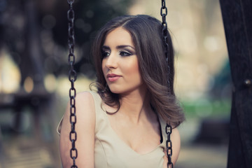 Outdoor portrait of beautiful young woman. She siting on park swing and looking at side.