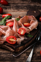 thin slices of prosciutto with mixed with basil, cherry tomatoes on wooden cutting board,vintage background