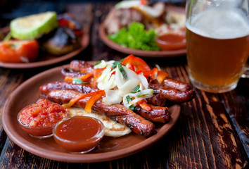 Barbecued pork or lamb meat sausages grill with salad, vegetables, glass of beer and sauce on clay dish. Vintage wooden table background.