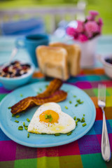 Tasty fried egg in the shape of a heart with bacon and toast on table in the garden.