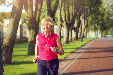Elderly woman running with headphones and smartwatches in the park in evening sunset