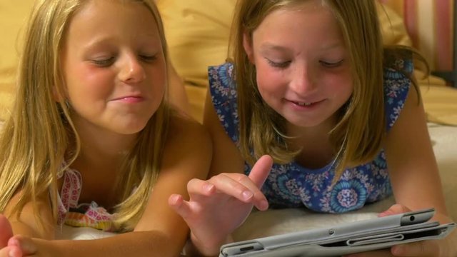 Two little girls lying on a bed are happily entertained by playing a game on a tablet pc.
