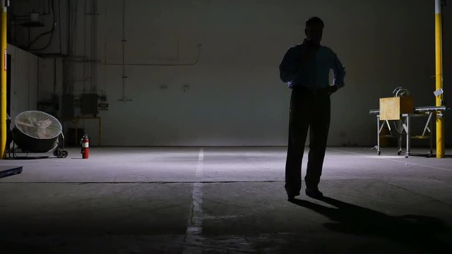A man on a cell phone pacing in the lone light of a large dark empty warehouse.