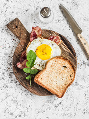 Toast with fried egg, bacon and arugula on the wooden cutting board. Delicious breakfast. On a light background, top view