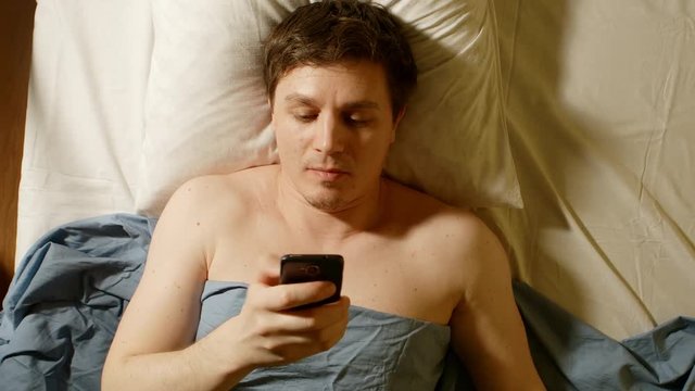 Caucasian young man types by mobile phone in a bed