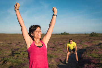 Smiling runner woman rising her arms after wining trail running training race. Couple competitively while preparing marathon or triathlon competition.