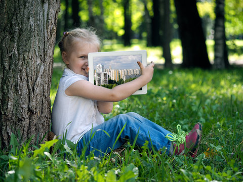 Little girl on the grass with tablet. Happy kid shows photos of the city on the tablet