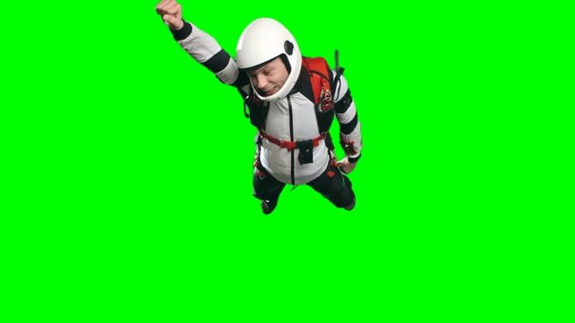 Slow motion shot of young man in white helmet and parachuting gear with one arm forward, doing superman flying pose and smiling and in free fall , green background, extreme sports chroma key