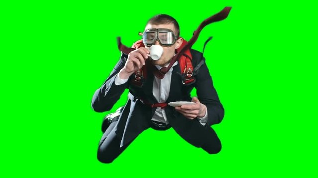 Calm and relaxed young businessman flying in free fall drinking coffee, wearing formal suit, aviator mask and parachute on his back, chroma key against green screen background