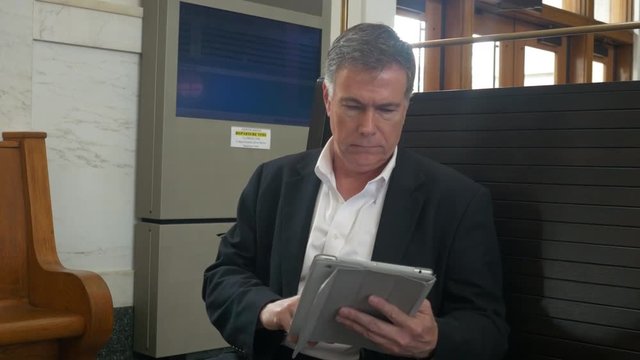 A mature businessman sitting on a large high backed bench in a historic train station using a tablet pc.
