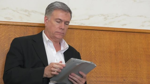 A mature businessman sitting on a large high backed bench typical of a vintage train station closes his tablet pc and looks around.