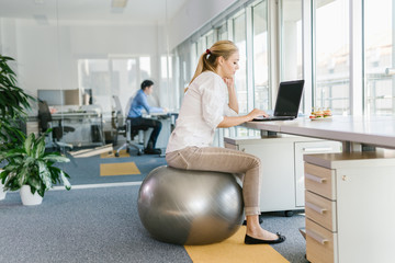 Business woman working on laptop in office while sitting on pilates ball