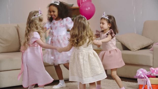 Four little girls dressed in pink dresses and tiaras dancing in circle and then one of them falling on the floor