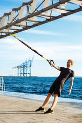 Young sportive man training with trx near sea in the morning.