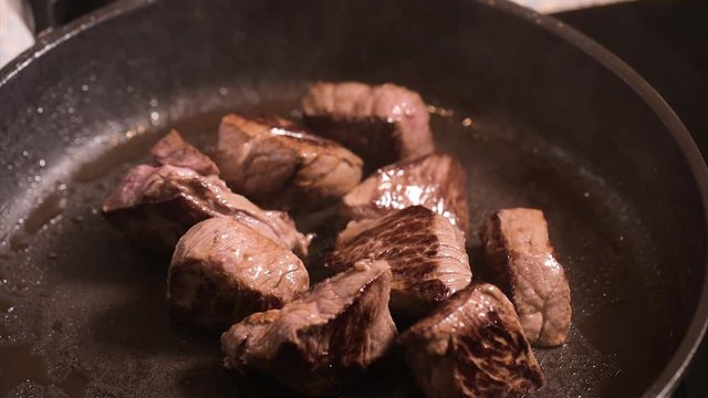 Frying beef sirloin in a hot pan. Turning pieces of  beef meat - close-up on top of a kitchen tile. Cook with fork preparing meat.