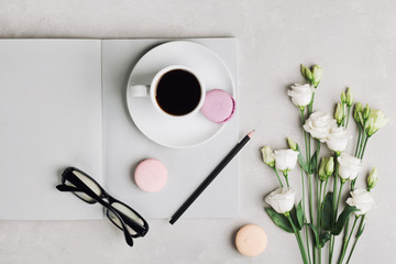 Morning cup of coffee, empty notebook, pencil, glasses, white flowers and cake macaron on light table top view. Beautiful breakfast. Flat lay.
