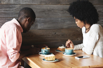 Fototapeta na wymiar Two black co-workers having launch together during coffee break. Young African man in glasses using smartphone while sitting at cafe with his dark-skinned girlfriend drinking tea and eating cakes