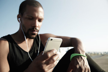 Portrait of pensive handsome dark-skinned man with little beard listening to music on his smartphone under blue sky. Lonely guy plunging into melodies and enjoying time spent alone with his thoughts.