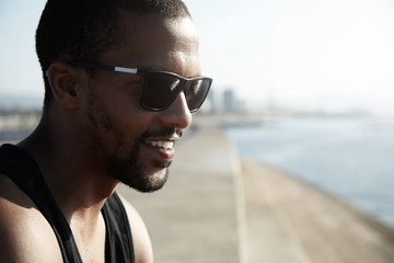 Charismatic dark-skinned guy in trendy sunglasses standing in morning sun admiring the beginning of day, smiling remembering something funny, against blurred background of big city landscape