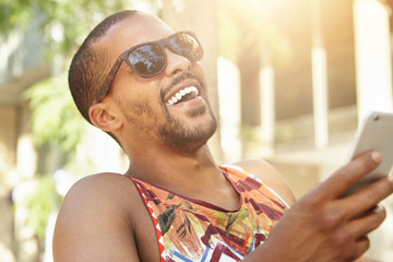 Human emotions and feelings. Cute charismatic dark-skinned man holding smart phone, laughing out loud after receiving hilarious picture from best friend, against background of summer city landscape