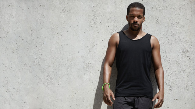 Handsome dark-skinned jogger with fit athletic arms dressed in black sleeveless shirt and shorts, looking at camera with serious and confident expression on his face standing by gray wall outdoors