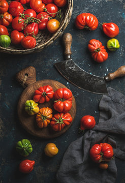 Fresh colorful ripe Fall heirloom tomatoes in basket and on wooden board, herb chopper knife for cooking over grunge dark plywood background, top view. Harvest vegetable cooking conception.