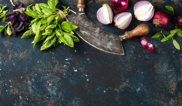 Healthy food cooking background. Fresh green and purple basil leaves, red onions and garlic with herb chopper knife over grunge dark blue painted plywood texture. Top view, copy space