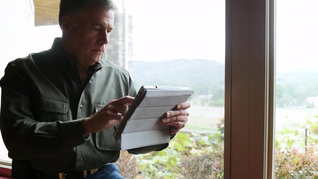 A contented mature man situated next to a window uses a digital tablet.