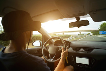 Naklejka premium Traveler in panama hat drives his car on interurban road during sunset, one hand on steering wheel and sun flares on windscreen