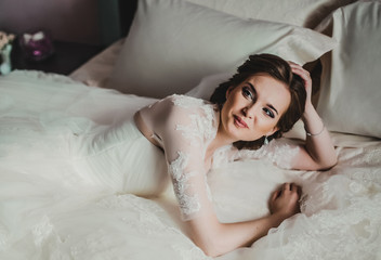 Portrait of beautiful bride with fashion veil posing on bed at wedding morning. Makeup. Brunette girl with long wavy hair styling. Wedding dress.