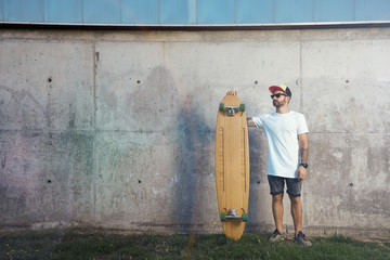 Young man wearing blank white t-shirt, baseball hat, shorts and sneakers standing with a wooden longboard next to a gray concrete wall