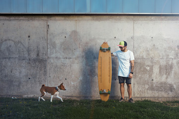 Bearded and tattooed longboarder standing next to a concrete wall looking at an approaching brown and white basenji dog
