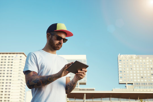 Focused young man with a beard and tattoos wearing an unlabeled white t-shirt looking at his tablet in a city