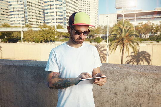 Focused bearded and tattooed young white man wearing a plain t-shirt and a baseball cap looking at his tablet in a southern city